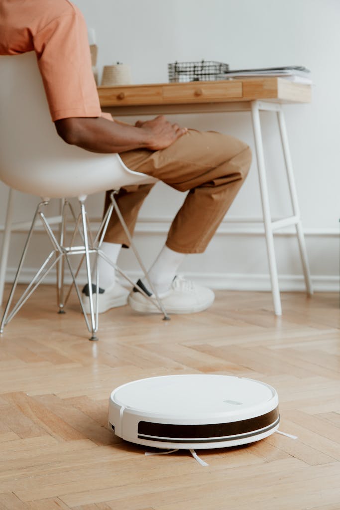A man in casual attire sits as a robotic vacuum cleans the wooden floor.