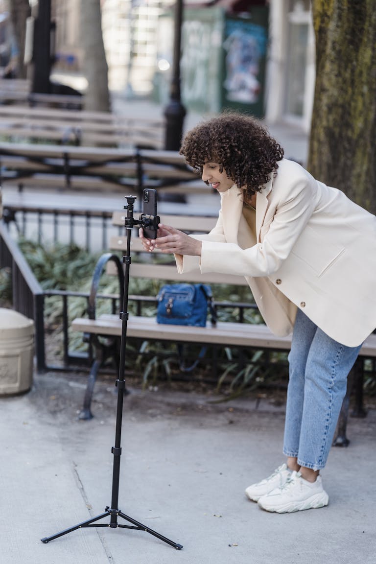 A young woman with curly hair uses a smartphone on a tripod in an urban park.