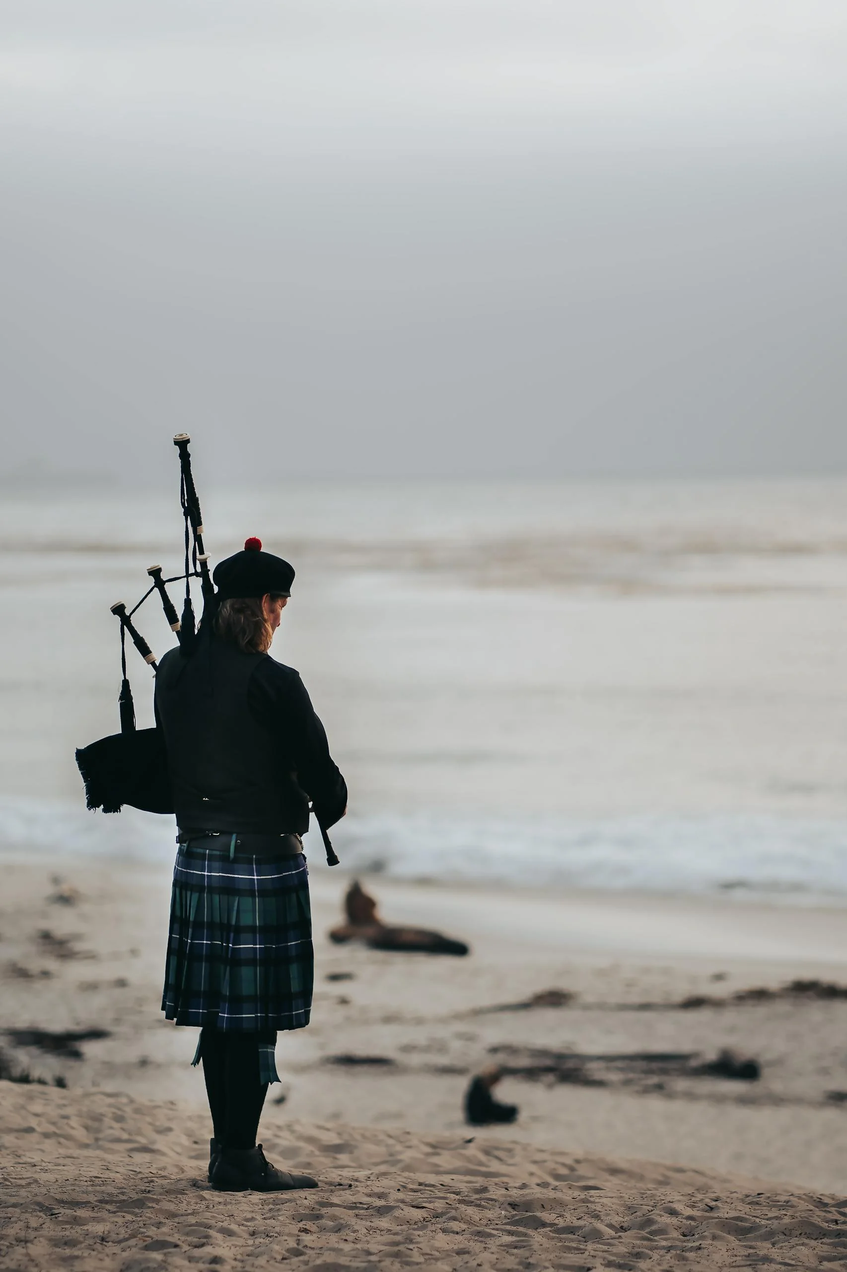 A bagpiper in traditional Scottish attire plays by the foggy shore of Carmel-by-the-Sea, creating a serene atmosphere.