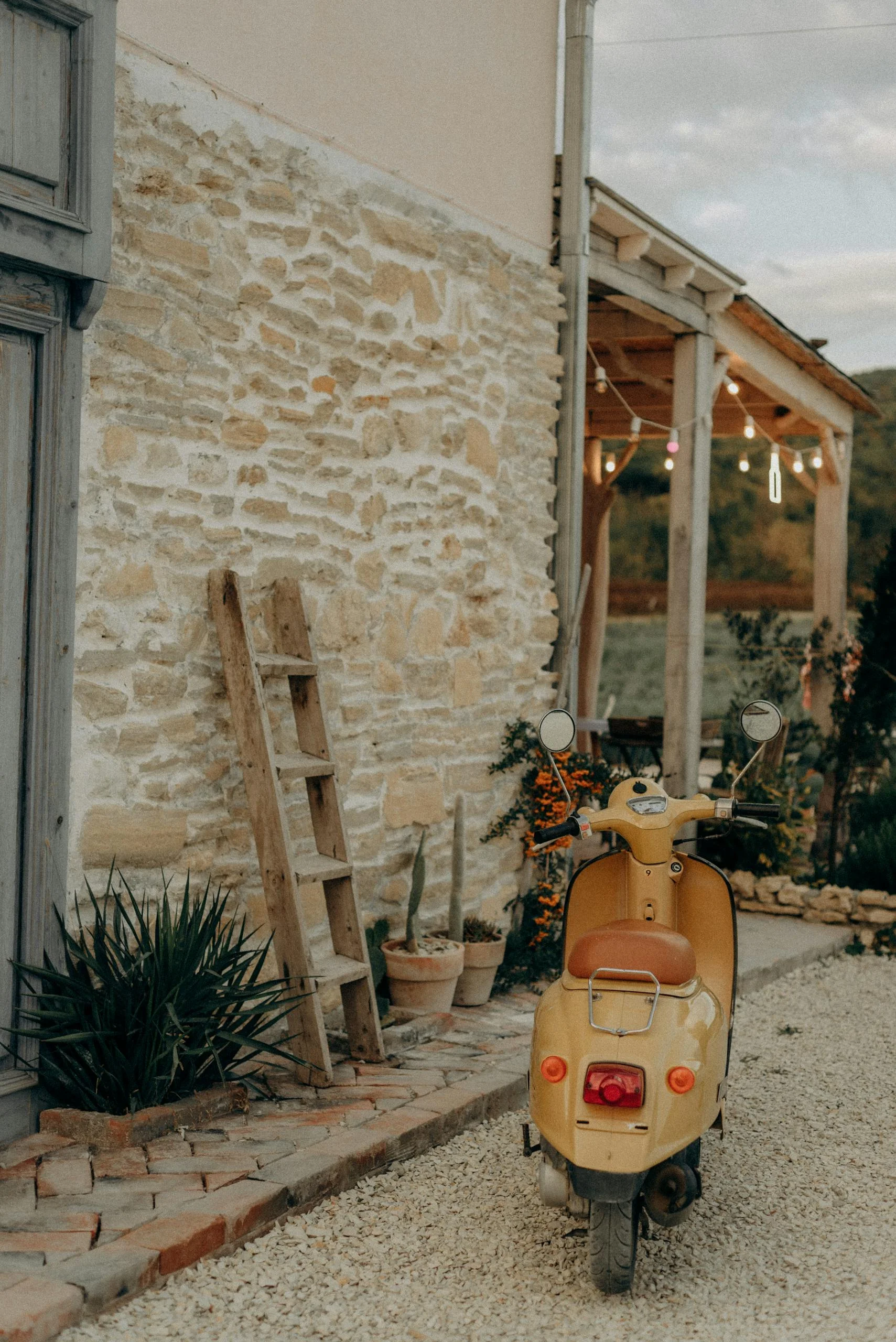 Charming scene of a vintage yellow scooter parked by a rustic stone wall with a wooden ladder and plants.