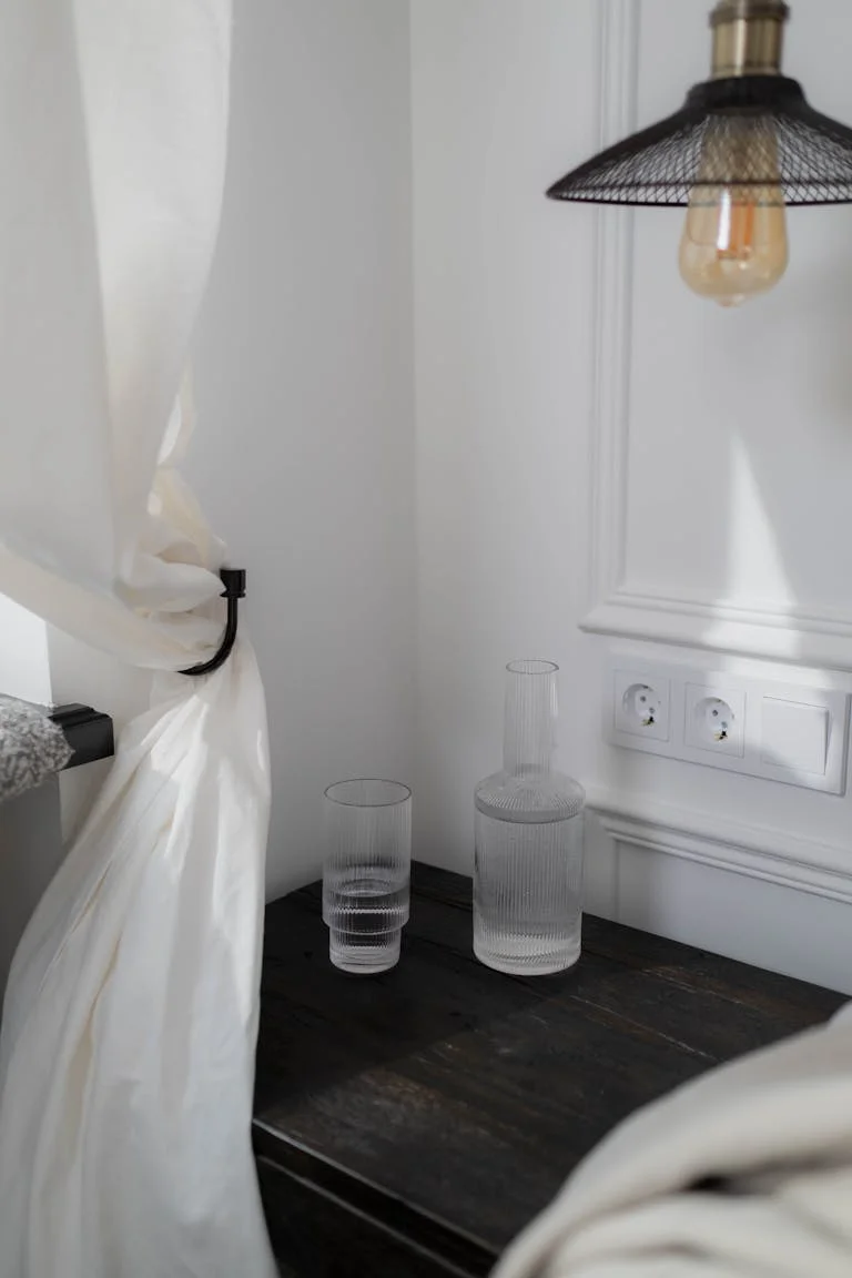 Stylish bedroom corner featuring a wooden table with glass bottles, soft white curtain, and hanging lamp.