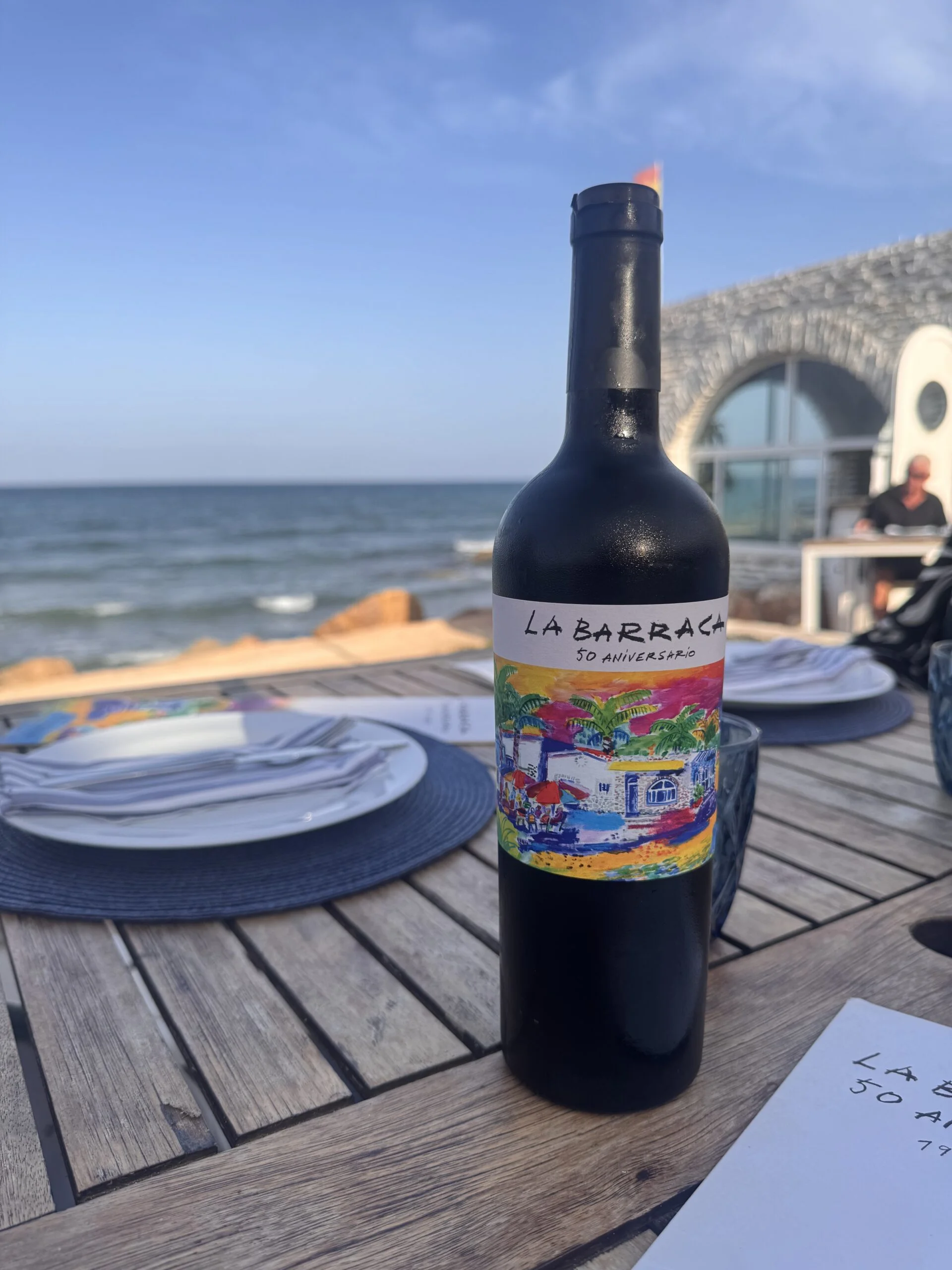 La Barraca 50th anniversary wine bottle on a wooden table overlooking the Mediterranean at Campoamor’s iconic beachfront restaurant. | elle blonde Luxury Lifestyle Destination Blog