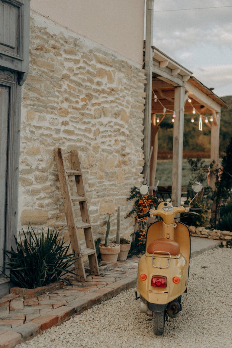 Charming scene of a vintage yellow scooter parked by a rustic stone wall with a wooden ladder and plants.