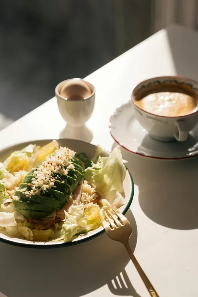 4 Simple Fit Body Tips To Help You Get Ready For Summer 3 A sunlit breakfast scene featuring a fresh avocado and shredded cheese salad on a bed of lettuce, with a soft-boiled egg and a cup of coffee on a white table. | Healthy Living & Wellness | Elle Blonde Luxury Lifestyle Destination Blog