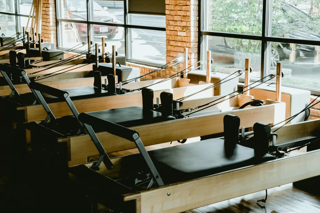 A row of wooden Reformer Pilates machines in a sunlit studio with large windows and exposed brick walls, reflecting a serene space for mindful movement and wellness. | Reformer Pilates & Healthy Wellbeing | Elle Blonde Luxury Lifestyle Destination Blog