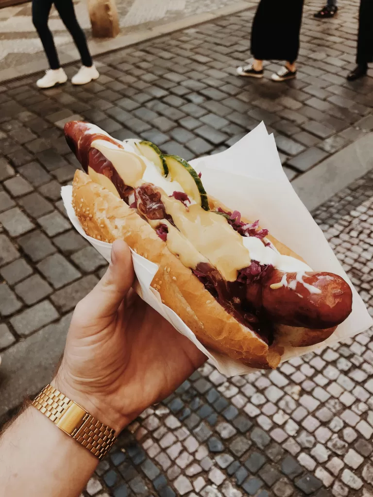 How To Plan An Unforgettable Stag Do In Prague 5 A hand holding a Czech street food hot dog topped with mustard, onions, and pickles over cobbled streets in Prague’s Old Town. czech street food for stag do in prague | best local eats in prague for bachelor parties | elle blonde luxury lifestyle travel blog