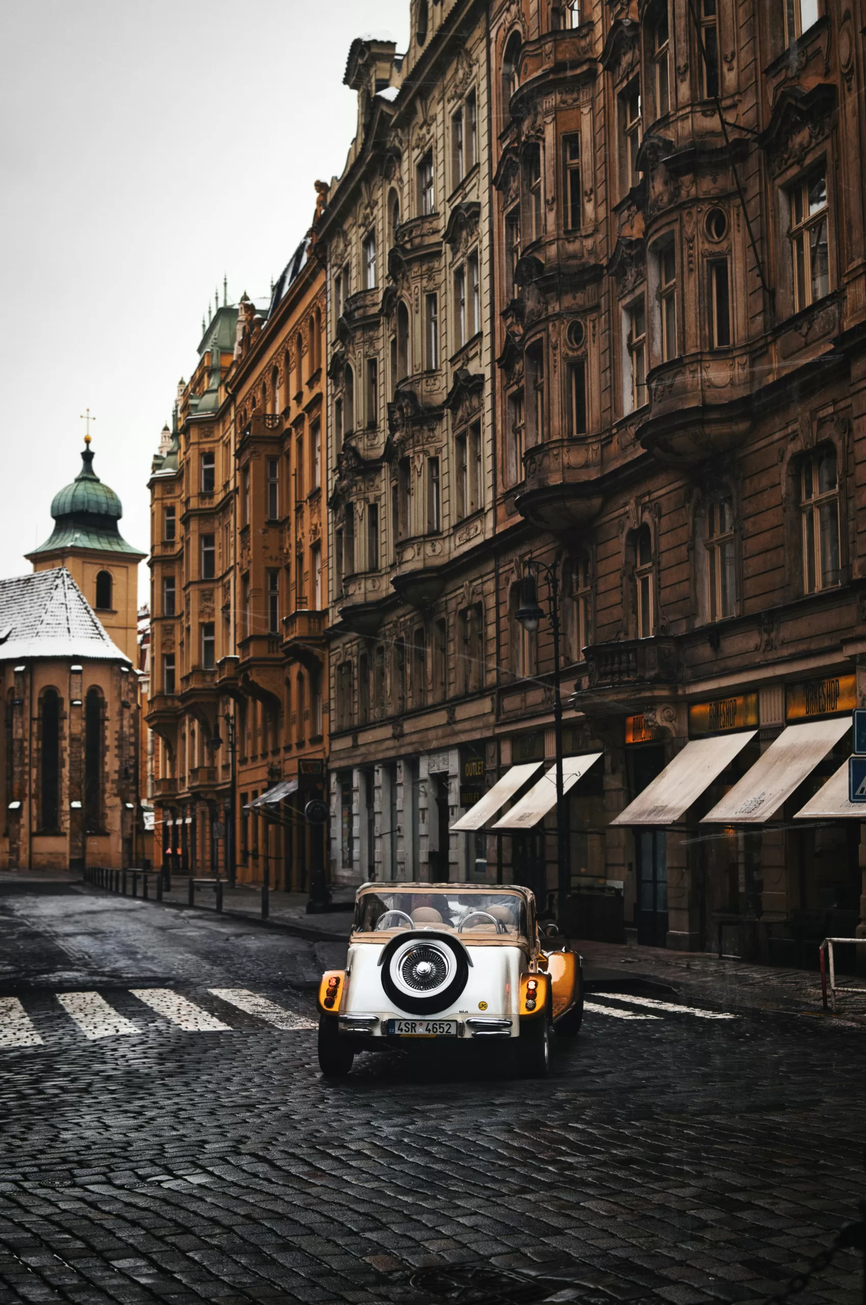 A vintage convertible car driving through Prague’s historic old town streets, lined with Baroque buildings on a winter morning. vintage car tour in prague | stag weekend activities with european flair | elle blonde travel tips for luxury city breaks