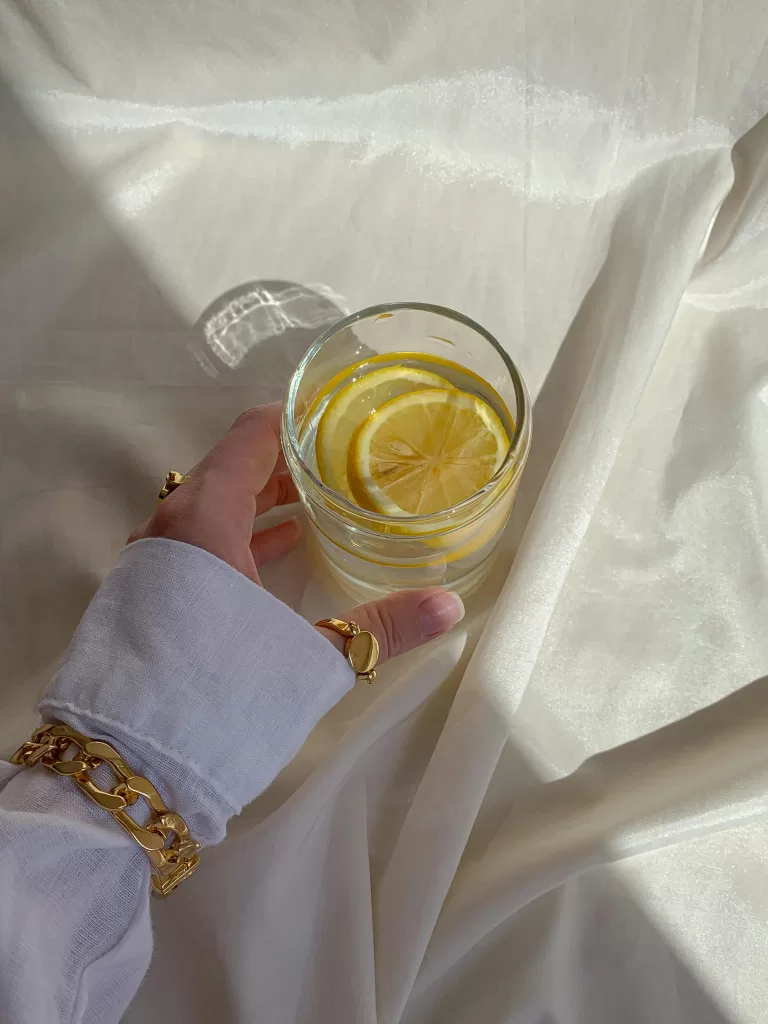 5 Restorative Techniques to Recharge After a Long Flight 1 A refreshing glass of lemon water held delicately over soft, cream satin bedding, accessorised with gold jewellery — evoking effortless elegance in a sunlit luxury resort suite. | 5 Restorative Techniques to Recharge After a Long Flight | Elle Blonde Luxury Lifestyle Destination Blog