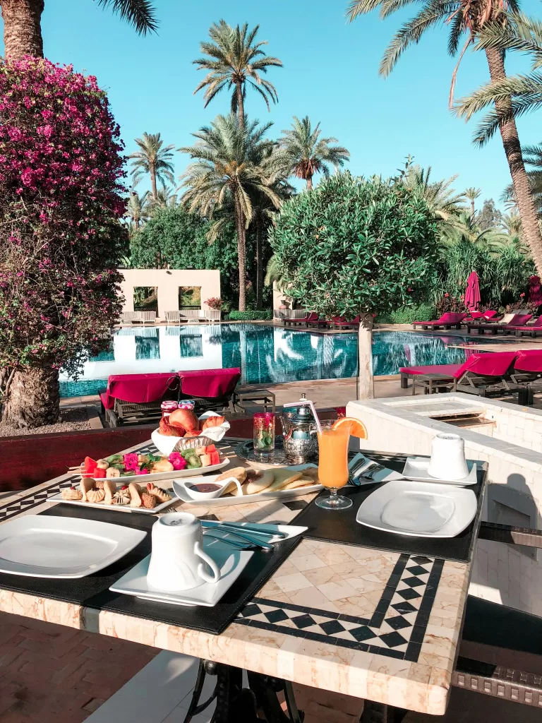 How to Make Business Travel Feel Like a Relaxing Getaway 6 Luxurious outdoor breakfast setup by a serene palm-lined pool in a tropical resort, complete with fresh fruit, pastries, and vibrant cocktails. | Travel Tips | Elle Blonde Luxury Lifestyle Destination Blog