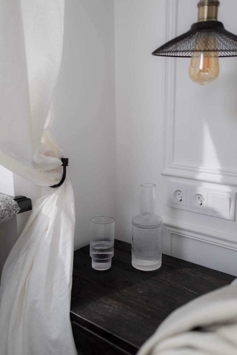 Stylish bedroom corner featuring a wooden table with glass bottles, soft white curtain, and hanging lamp.